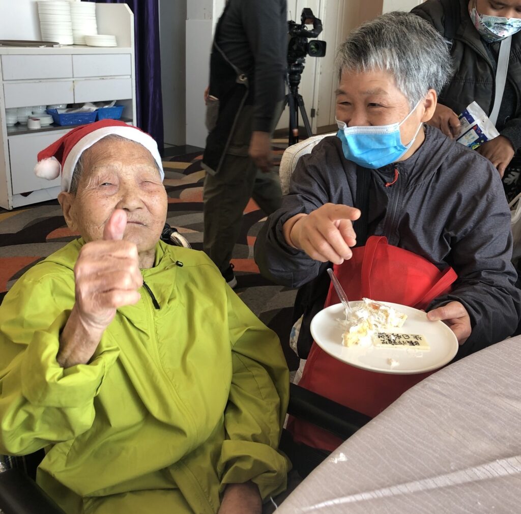 A photo of an elderly in a wheelchair and one woman feeding her, with a happy face and a good hand sign
一名輪椅人士及一名女士正在餵食，並展露笑容的合照。
